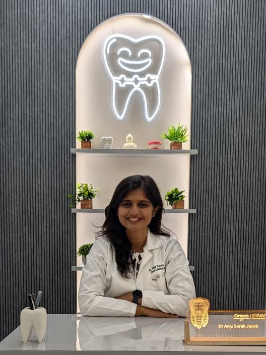Dr. Anju Sarah Jacob at her desk in the studio, under the lit arch with the practice's tooth in it
